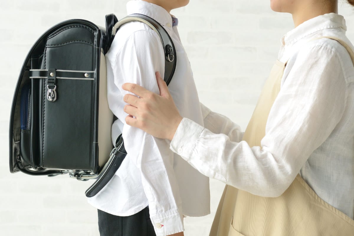 ランドセルを背負った子どもに寄り添い、そっと肩に手を添えて励ましている保護者の姿。学校への行き渋りや不安を抱える子どもへのサポートを象徴するシーン。