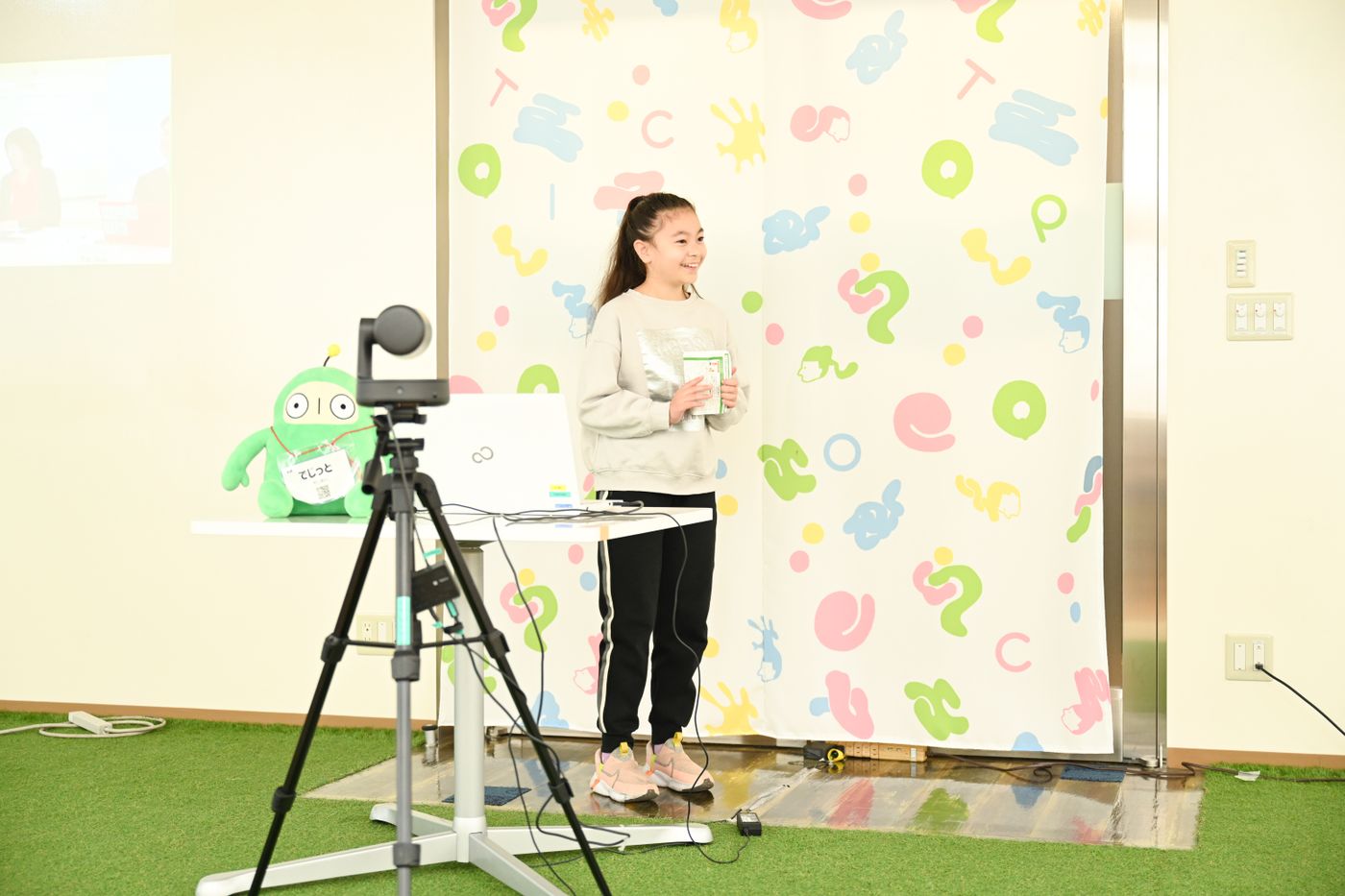 カラフルでポップな背景を背に、プレゼンテーションが行われている様子。三脚に取り付けられたカメラやノートパソコン、イベントのマスコットキャラクターがディスプレイに並ぶ。