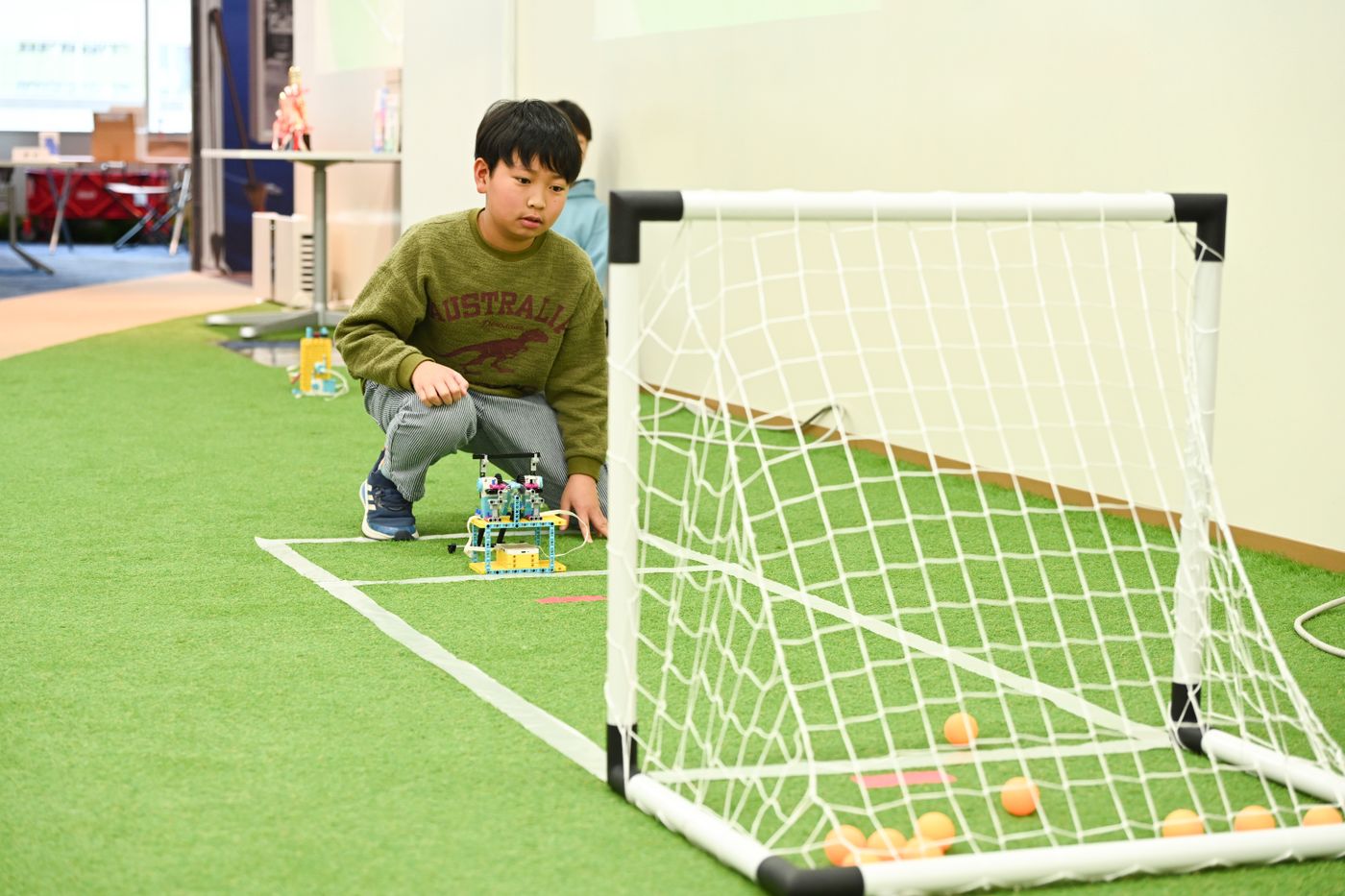 ロボットを操作してゴールにピンポン玉を入れる競技に取り組む様子。人工芝のフィールドに設置されたゴールネットを目指し、手作りのロボットが活躍している場面。