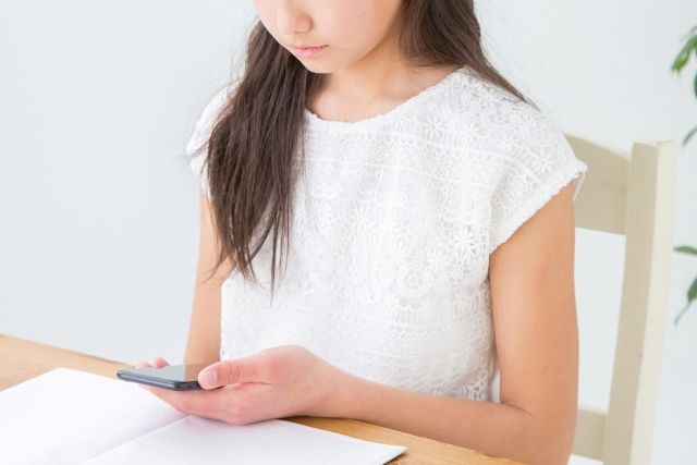 テーブルの前に座る人物がスマートフォンを操作している様子を捉えた画像である。白いレースのトップスを着ており、テーブルの上には開かれたノートや本が置かれている。