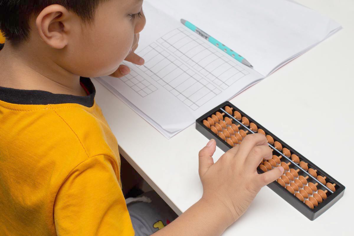 子どもが算数の勉強をしている様子を捉えた画像である。テーブルの上には教科書のような大きなノートが広げられ、その横には算盤（そろばん）が置かれている。子どもは右手で算盤の玉を操作しており、学習に集中している様子が伺える。