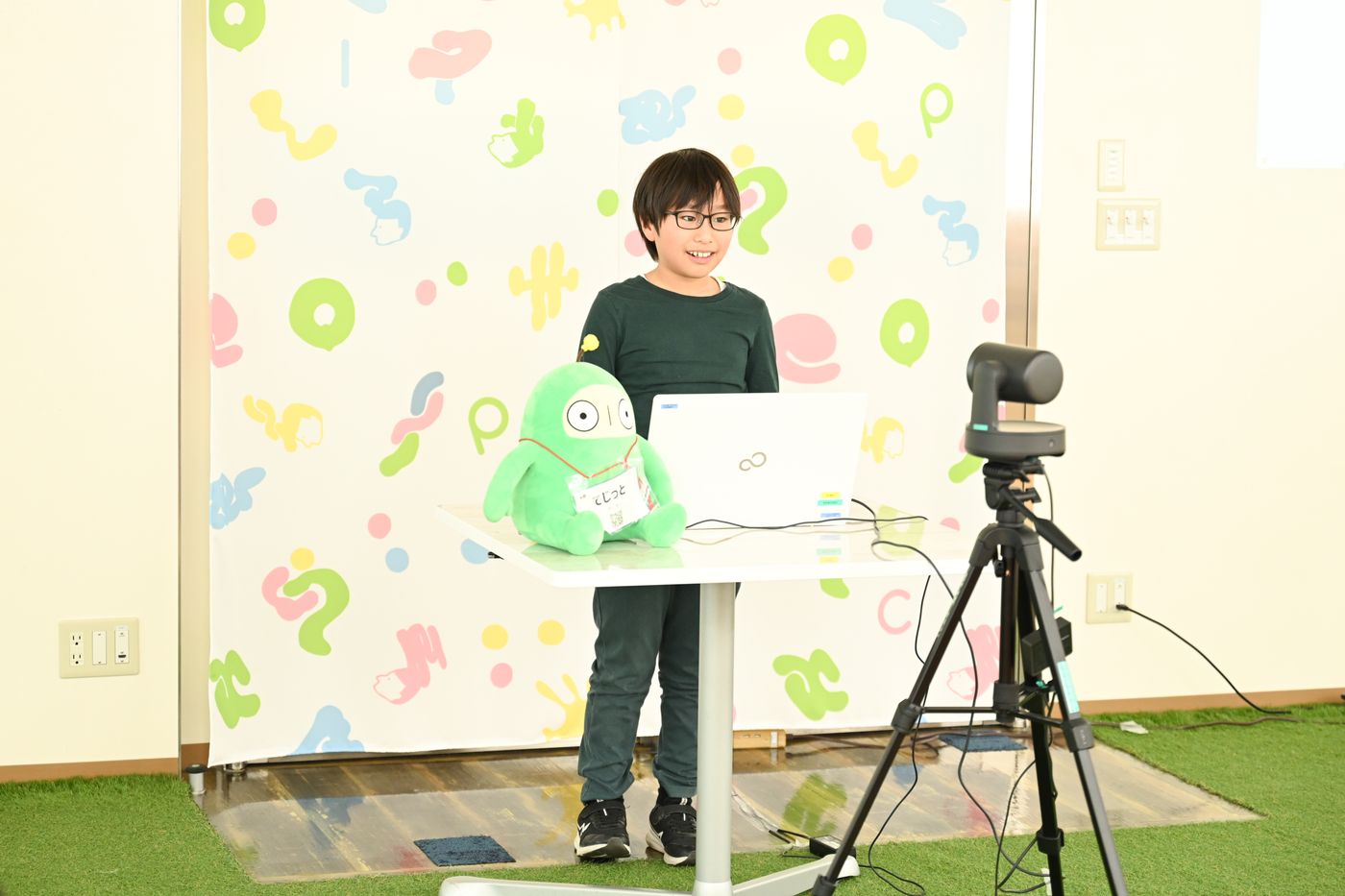 カラフルな背景を背に、机の上に置かれたノートパソコンとキャラクターぬいぐるみ『てしつと』が配置されたプレゼンテーション会場の様子。三脚に設置されたカメラも使用されている。