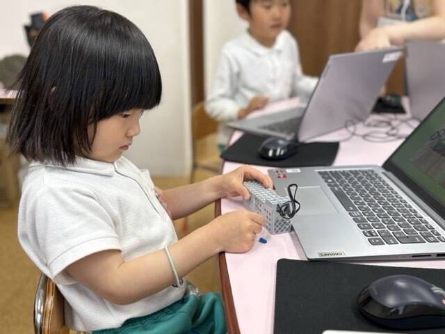 授業風景：女の子がパソコンの前に座り、ブロックを手に持っている