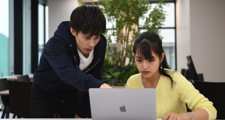 女性がPCに向けって座っており、男性が女性に対してPCに指差しながら教えている姿が映っている