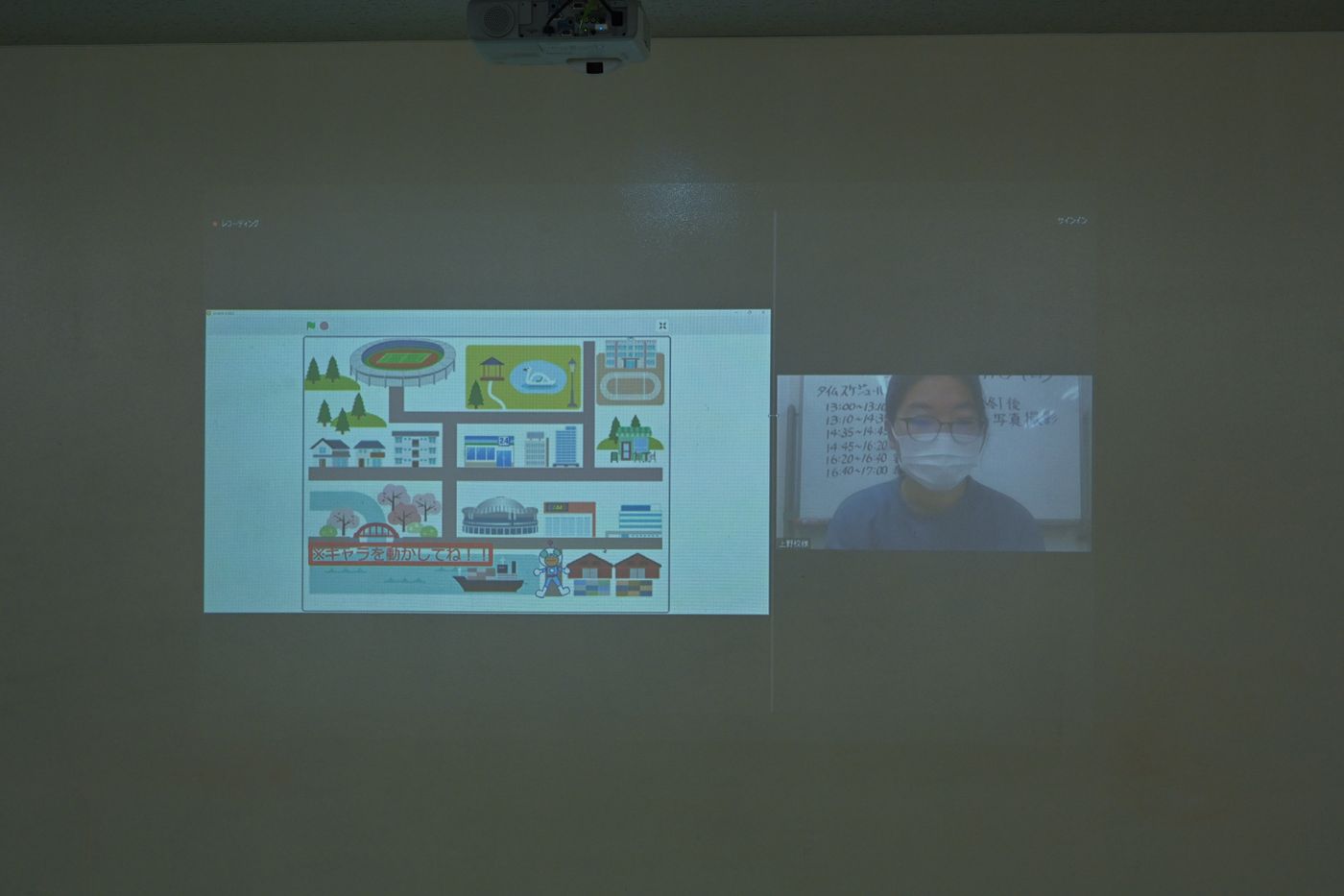 プロジェクターで映し出された画面には、イラストで描かれた街並みや施設が表示されており、別の画面にはタイムスケジュールが書かれたホワイトボードが映っている。教育やプレゼンテーション用途の場面を示している。