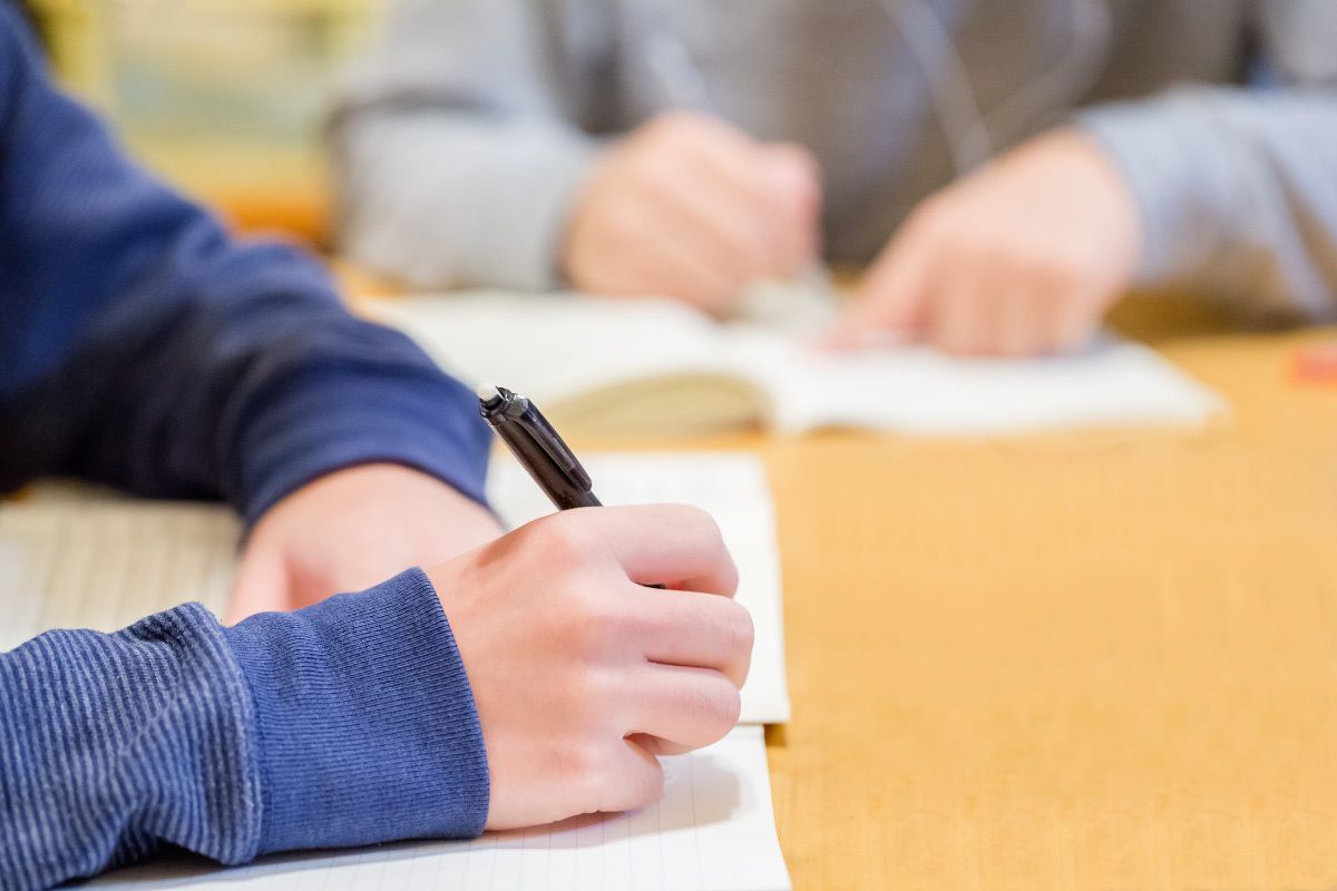 机に向かってノートにペンで書き込みをしている学生の手元。勉強や学習風景を表した場面で、背景には開いた本が見える。
