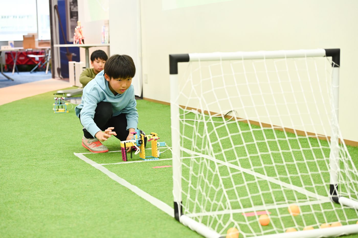 子どもがロボットを使ってピンポン玉をゴールに入れる競技に挑戦している様子。芝生のようなフィールドとゴールネットが設置された会場で、創造性を活かしたロボット操作が行われている。