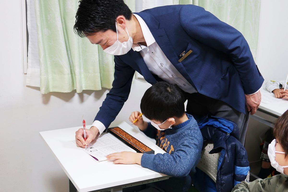 マスクを着用している男性が子供の机の後ろに立って男の子に教えている